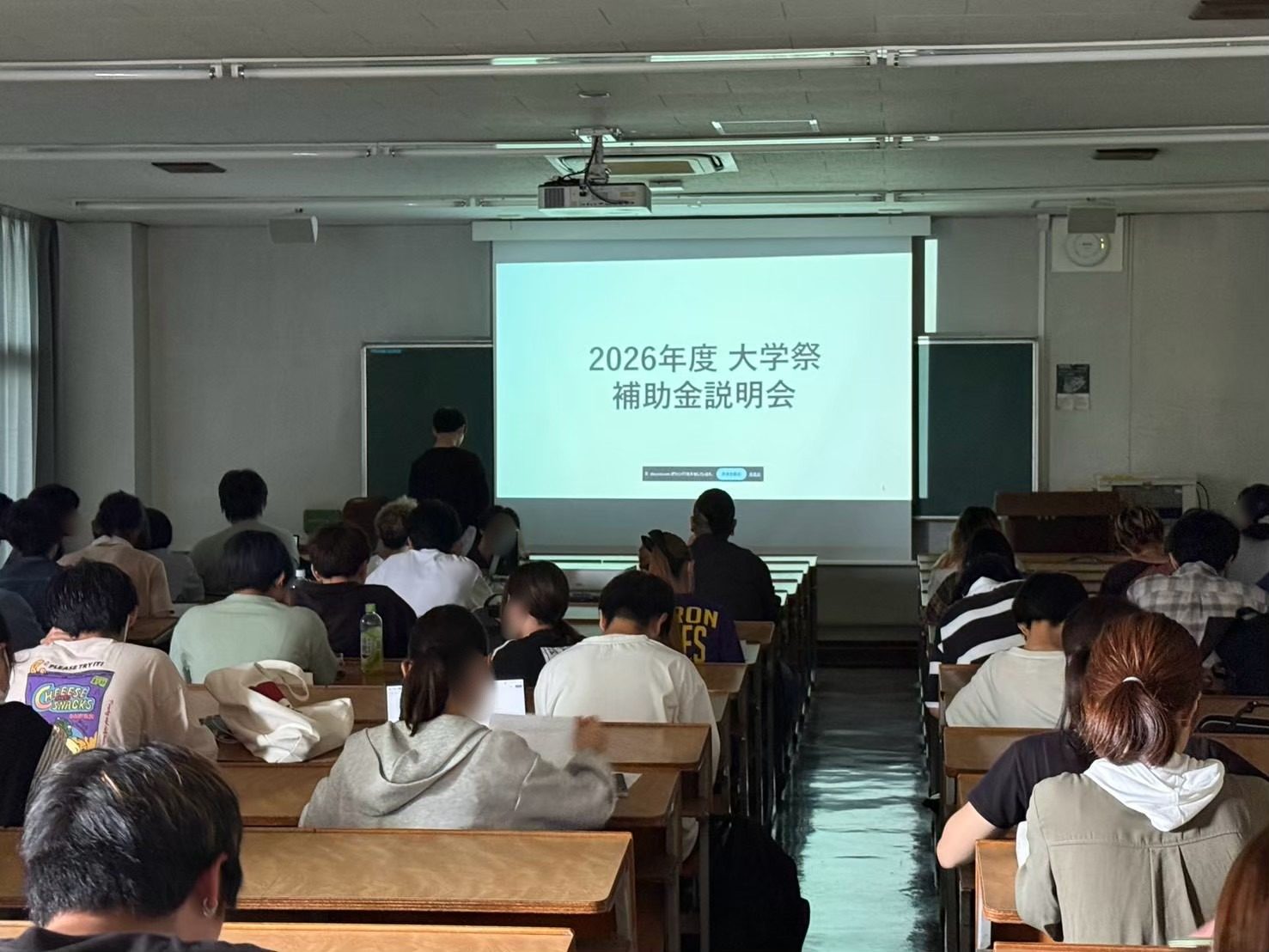 スクリーンに「2026年度 大学祭 補助金説明会」と映した教室と、席について始まりを待つ参加団体の学生