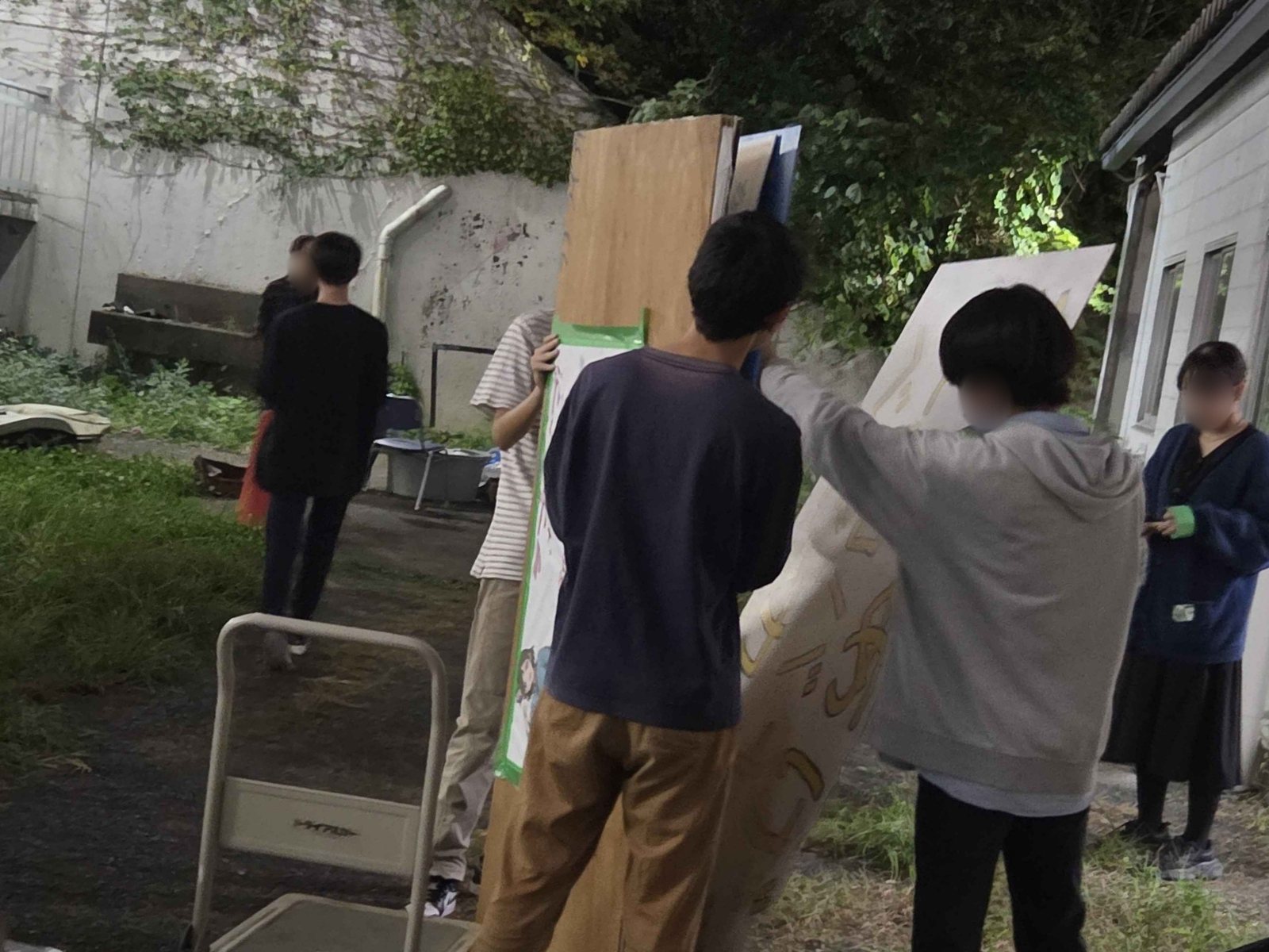暗くなった屋外で、大きな看板の板を数人で運ぶ実行委員