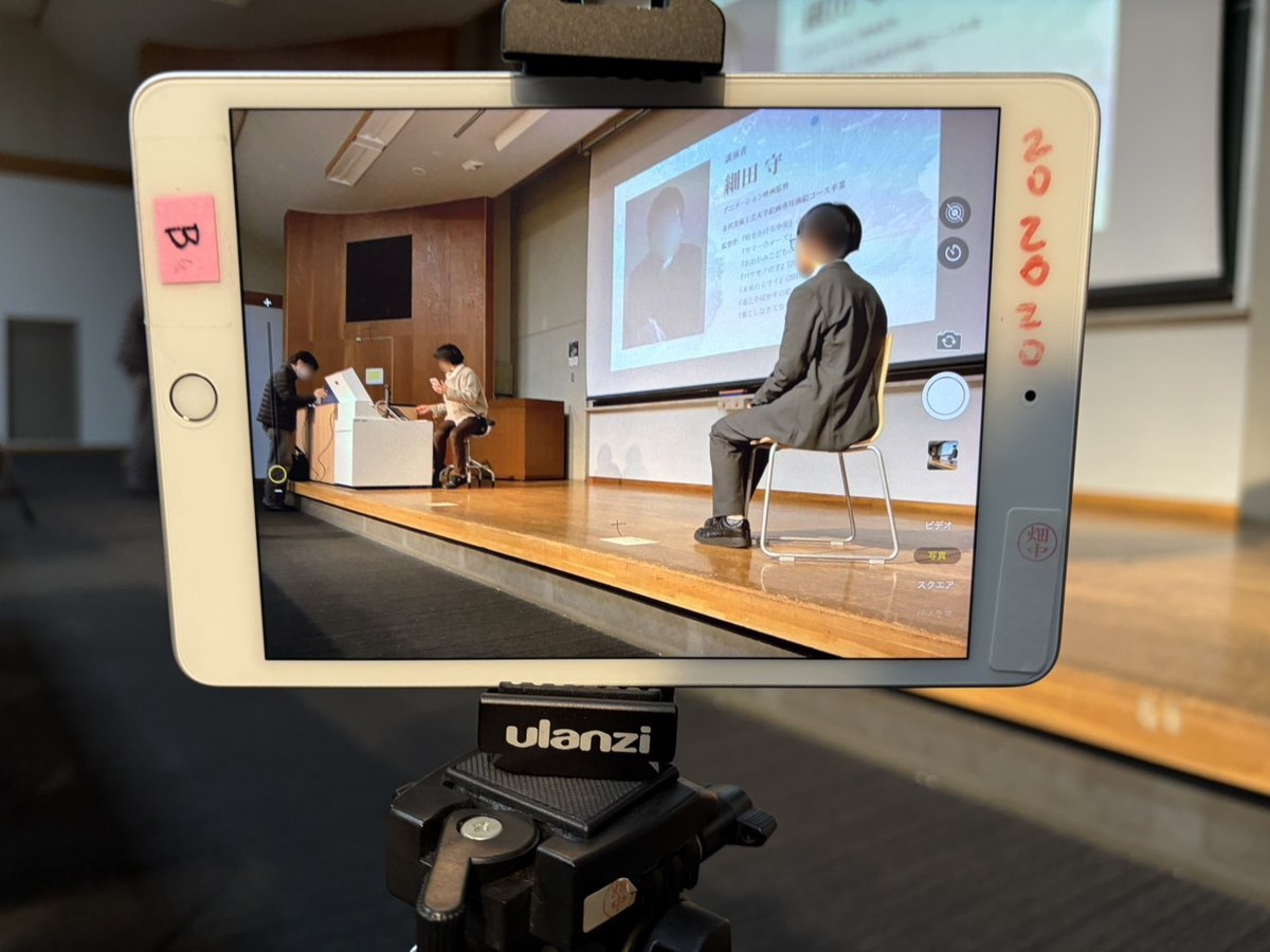 三脚に付けたタブレットの画面に、ステージの椅子に座るスタッフと、スクリーンに映る講演者「細田守」のスライドが映っている